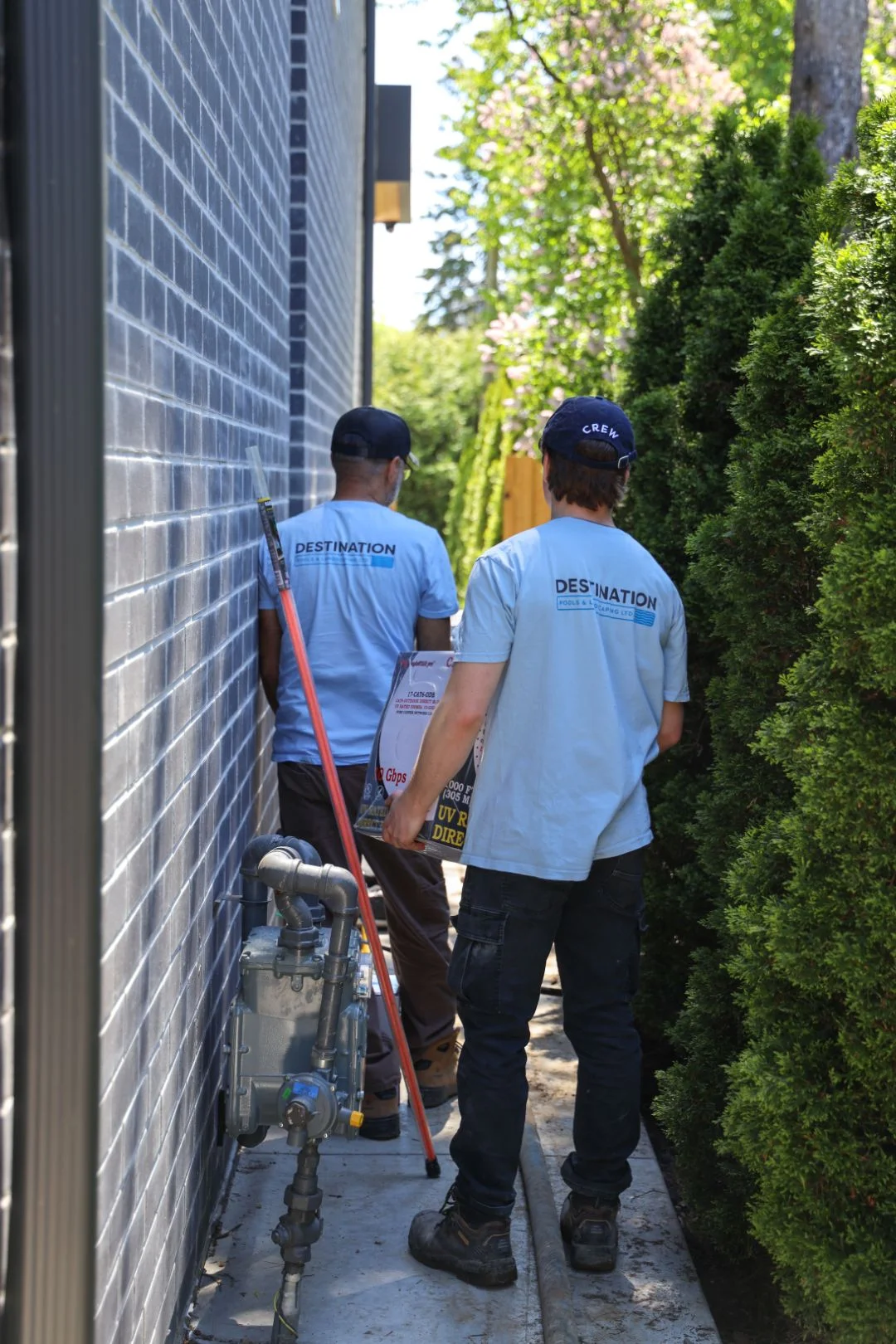 Two Destination Pools workers walking beside house to inspect gas service connection during backyard project