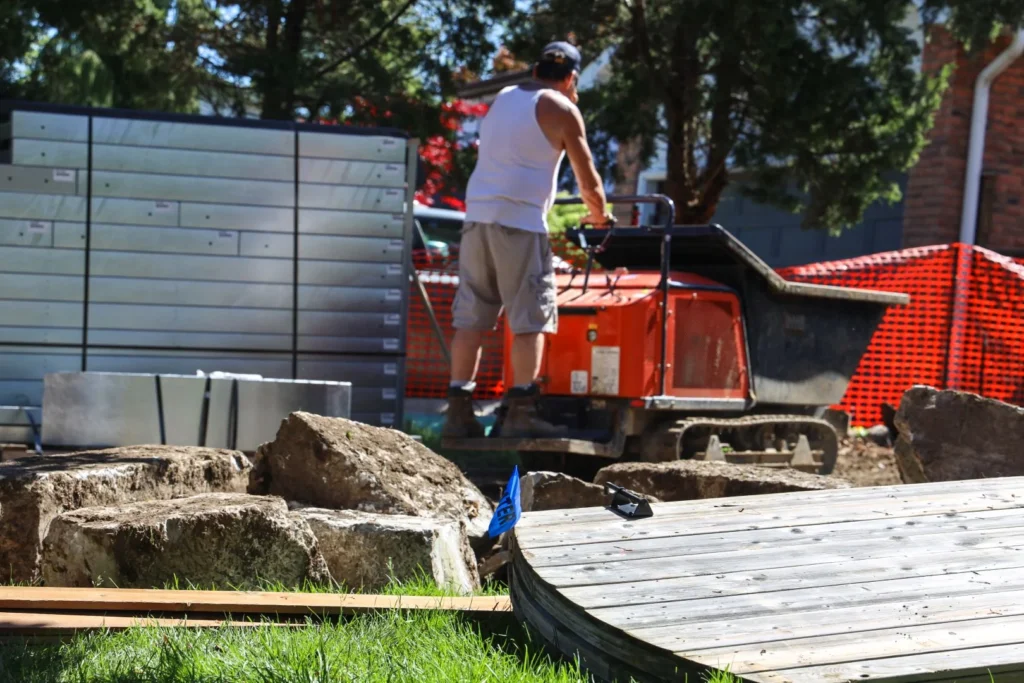 Worker operating Dingo compact track loader at backyard pool excavation site with stone materials